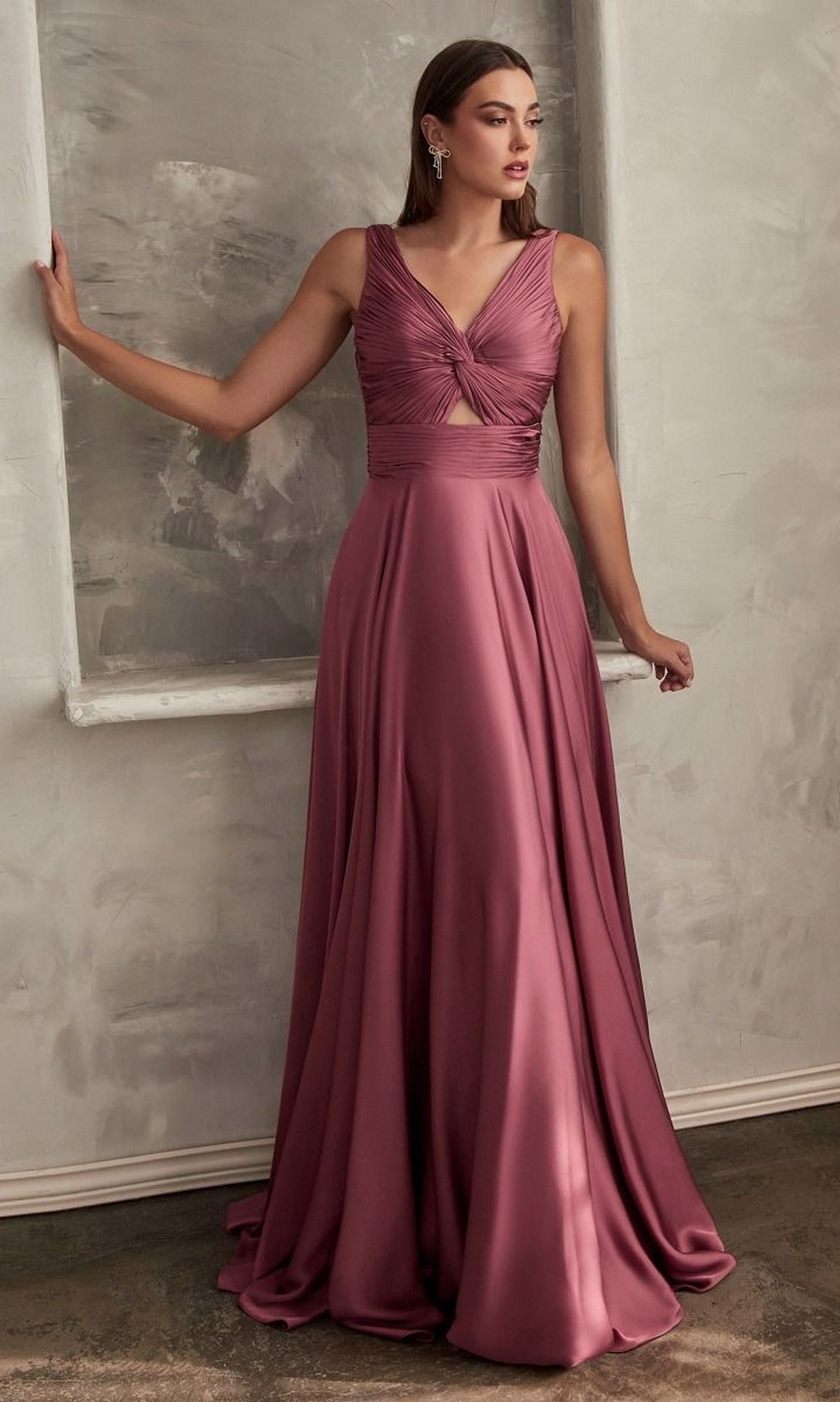 A woman poses indoors in the Twist-Front Long A-Line Cut-Out Prom Dress 7497—a sleeveless, floor-length mauve gown with a gathered bodice and front cutout—her arm gracefully resting on a frame.